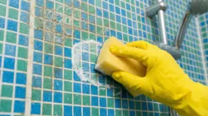 Mano con guanto giallo pulisce piastrelle a mosaico del bagno con una spugna